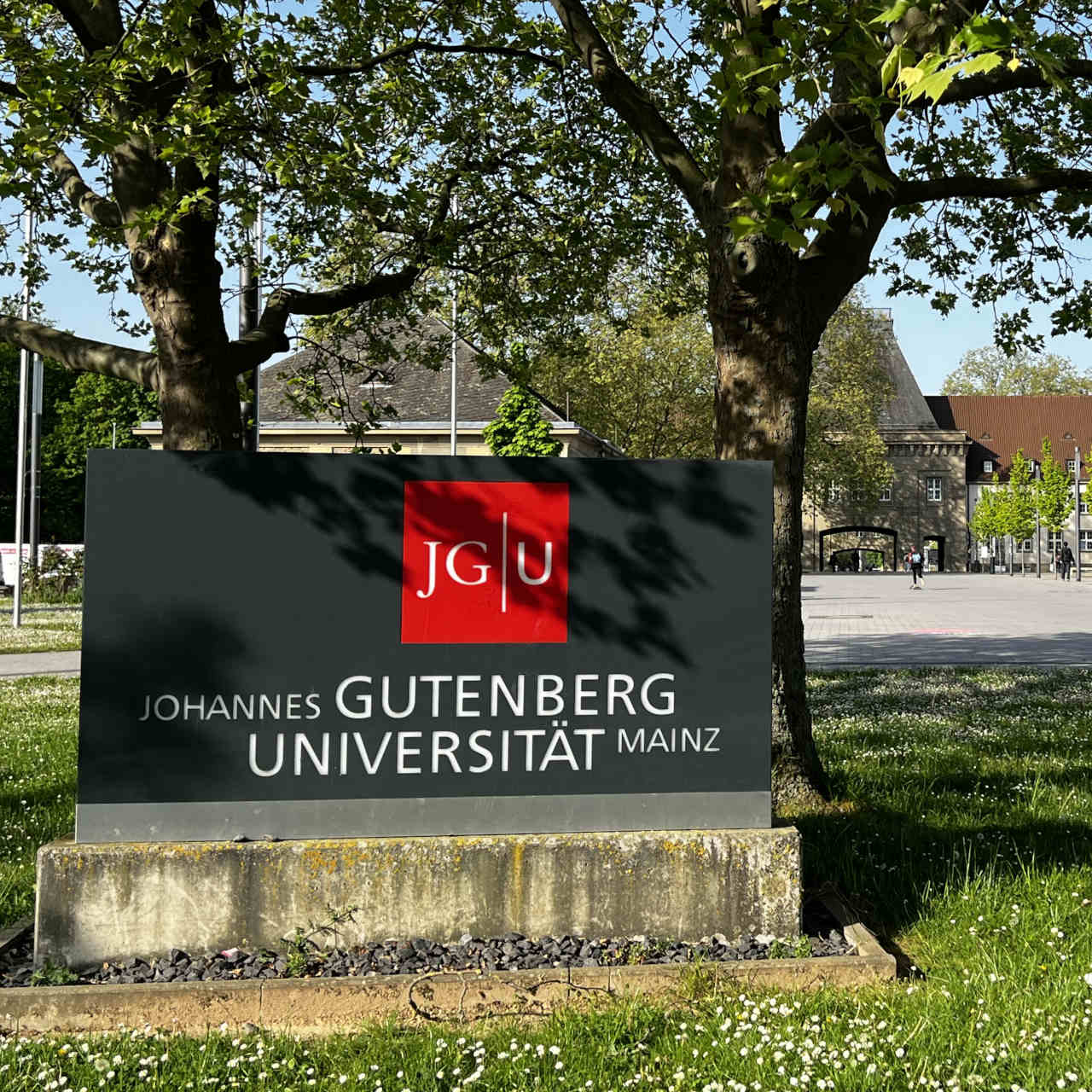 Das JGU-Schild am Eingang zur JGU von der Stadt aus. Im Hintergrund ist der markante Torbogen des Forums zu sehen.